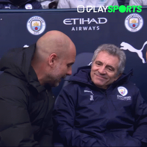 Two Men Are Sitting In Front Of A Manchester City Advertisement GIF