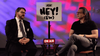 Two Men Are Sitting In Front Of A Sign That Says Hey ( Ew ) GIF