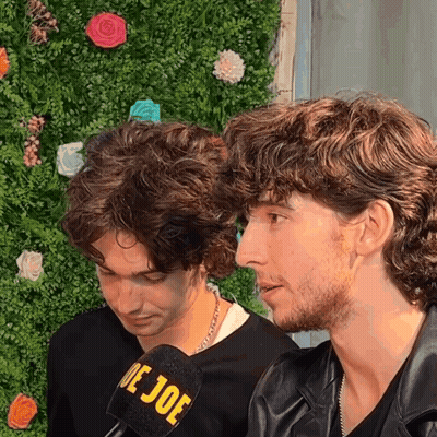 Two Men Are Sitting In Front Of A Wall With Flowers And A Microphone That Says De Joe GIF