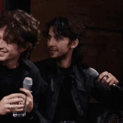 Two Men Are Sitting Next To Each Other Holding Microphones And Drinking . GIF