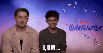 Two Men Are Sitting Next To Each Other In Front Of A Netflix Advertisement For Heartstopper GIF