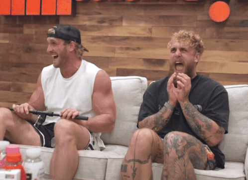 Two Men Are Sitting On A Couch Laughing And One Has A Tattoo On His Leg GIF