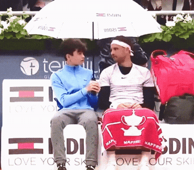Two Men Are Sitting Under An Umbrella In Front Of A Sign That Says Love Your Skin GIF
