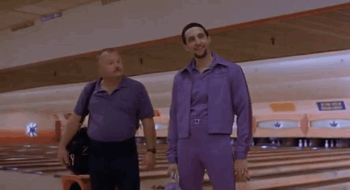 Two Men Are Standing In A Bowling Alley Wearing Purple Jumpsuits . GIF