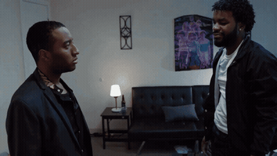Two Men Are Standing In A Living Room With A Purple Poster On The Wall GIF