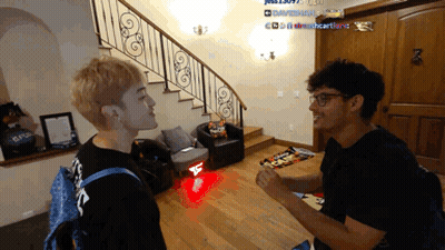 Two Men Are Standing In A Living Room With A Staircase In The Background And One Of Them Has A Backpack That Says Louis  GIF
