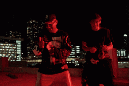 Two Men Are Standing In A Parking Lot At Night With A City In The Background GIF