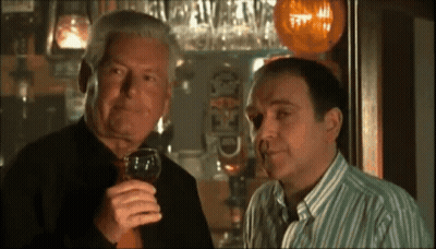 Two Men Are Standing In Front Of A Bar And One Is Holding A Glass Of Wine In His Right Hand GIF