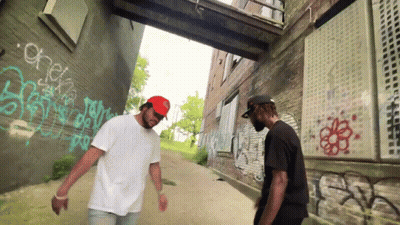 Two Men Are Standing In Front Of A Brick Wall With Graffiti On It Including One That Says Check GIF