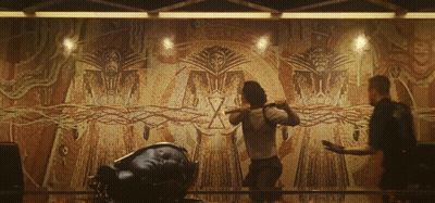 Two Men Are Standing In Front Of A Large Wall With A Drawing Of A Man With A Sword GIF