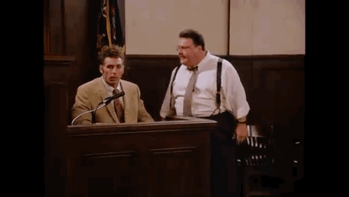 Two Men Are Standing In Front Of A Podium In A Courtroom Talking Into Microphones . GIF