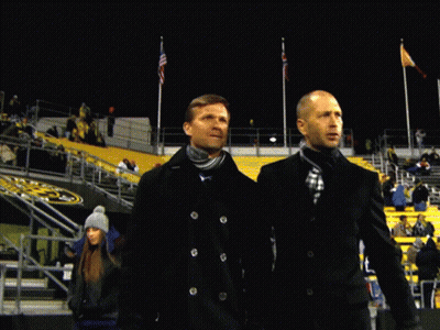 Two Men Are Standing In Front Of A Stadium With A Yellow Sign That Says ' T ' On It GIF