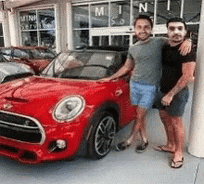 Two Men Are Standing Next To A Red Car In Front Of A Car Dealership . GIF