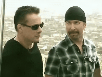 Two Men Are Standing Next To Each Other And Talking . One Of The Men Is Wearing Sunglasses And A Beanie . GIF