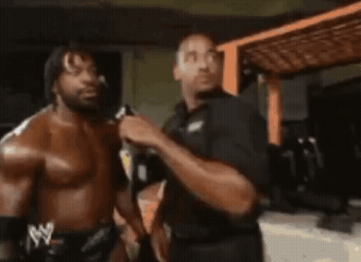 Two Men Are Standing Next To Each Other In A Room . One Of The Men Is Wearing A Shirtless Wrestling Outfit . GIF