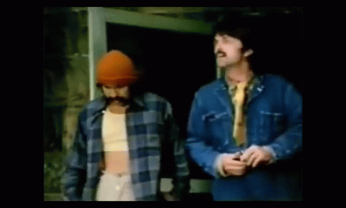 Two Men Are Standing Next To Each Other In Front Of A Door . One Of The Men Is Wearing A Hat And Tie . GIF