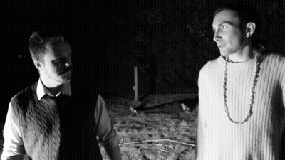 Two Men Are Standing Next To Each Other In The Dark And One Has A Necklace Around His Neck GIF