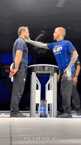 Two Men Are Standing Next To Each Other On A Stage And One Of Them Is Wearing A Blue Shirt That Says Slap League . GIF