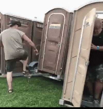 Two Men Are Standing Next To Portable Toilets In A Grassy Field . GIF