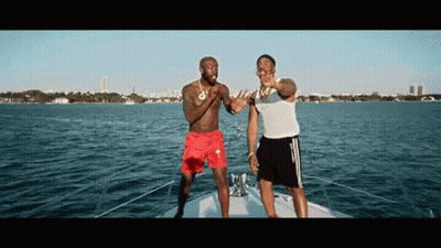 Two Men Are Standing On A Boat In The Water . One Of The Men Is Wearing Red Shorts . GIF