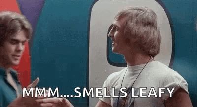 Two Men Are Talking To Each Other In Front Of A Sign That Says Smells Leafy . GIF