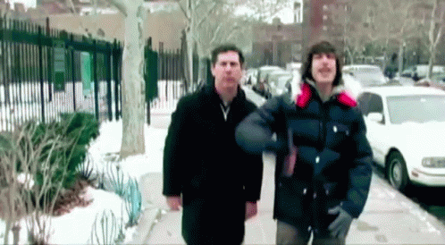 Two Men Are Walking Down A Snowy Sidewalk In Front Of Cars GIF