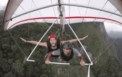 Two Men Flying Hang Glider Over Green Hillside GIF