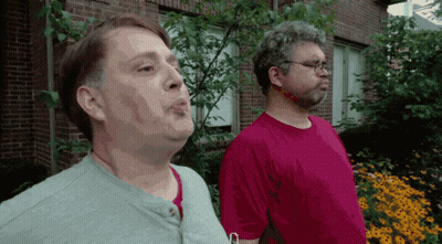 Two Men In Front Of Brick Building With Flowers GIF