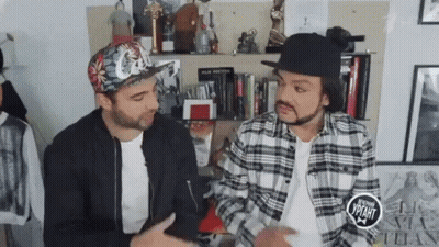 Two Men In Hats And Plaid Shirts Sitting By Bookshelf GIF