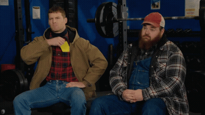 Two Men In Plaid Shirts And Overalls Are Sitting In A Gym One Is Holding A Yellow Card GIF