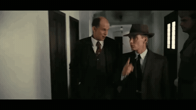 Two Men In Suits And Hats Are Standing Next To Each Other In A Hallway Talking To Each Other . GIF