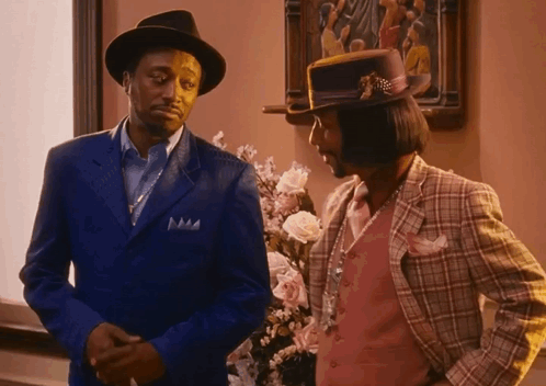 Two Men In Suits And Hats Are Standing Next To Each Other In Front Of Flowers GIF