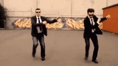 Two Men In Suits And Ties Are Dancing In A Parking Lot In Front Of A Graffiti Wall . GIF