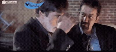 Two Men In Suits And Ties Are Talking In Front Of A Brick Wall With A Blue Sign That Says Pichuka GIF