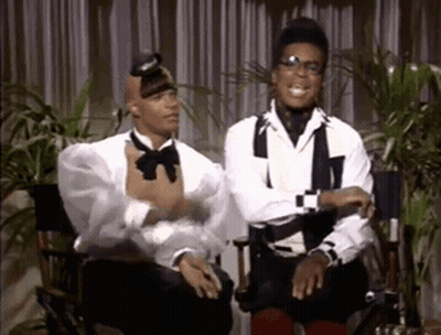 Two Men In Tuxedos Sitting In Director's Chairs Talking GIF