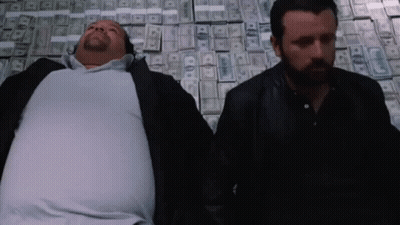 Two Men Laying On Top Of A Pile Of Money With One Of Them Having A Beard GIF