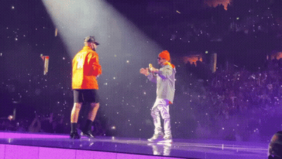 Two Men On A Stage With One Wearing An Orange Jacket With The Number 12 On It GIF