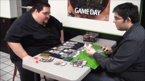 Two Men Playing A Game Of Cards With A Game Day Poster Behind Them GIF