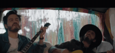 Two Men Playing Guitars In Van With Colorful Curtain GIF