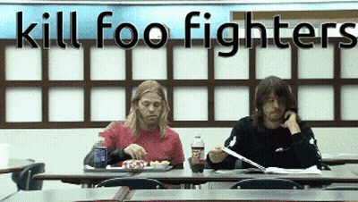 Two Men Sit At A Table In Front Of A Wall That Says Kill Foo Fighters On It GIF