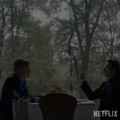 Two Men Sit At A Table With A Bottle Of Champagne And A Bucket Of Ice GIF