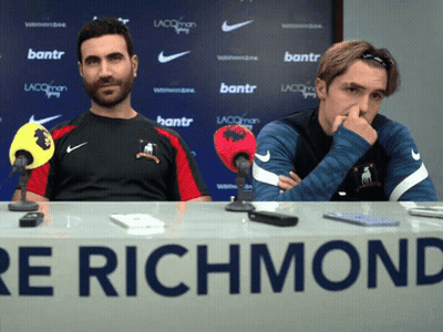 Two Men Sit At A Table With A Sign That Says Richmond GIF