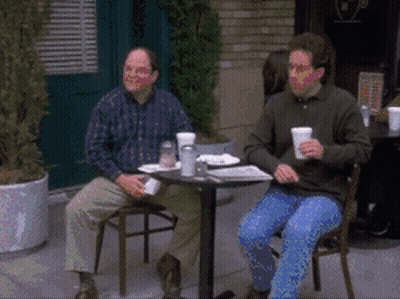 Two Men Sit At A Table With Cups In Their Hands GIF