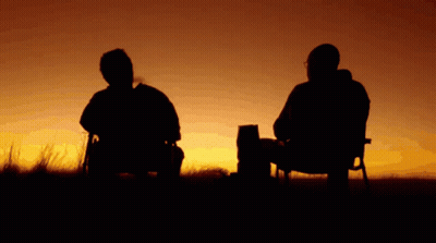 Two Men Sit In Chairs In Front Of A Sunset In A Field GIF