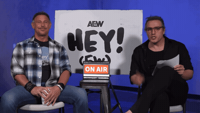 Two Men Sit In Front Of A Sign That Says " Hey " GIF