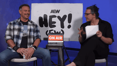 Two Men Sit In Front Of A Sign That Says Hey On Air GIF