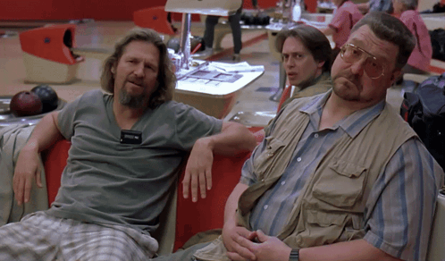 Two Men Sit On A Couch In A Bowling Alley With A Bowling Ball Behind Them GIF