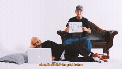 Two Men Sit On A Couch One Holding A Sign That Says Turn On The Printers Fellos GIF