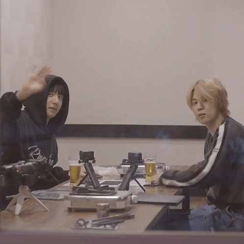 Two Men Sitting At A Table With A Camera And A Glass Of Beer One Of Them Has A Hoodie That Says Reign On It GIF