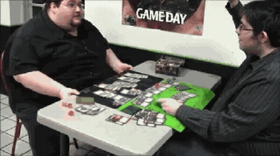 Two Men Sitting At A Table With A Game Day Poster Behind Them GIF